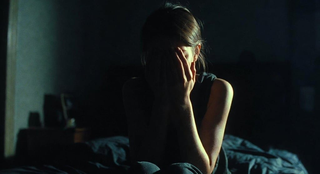 A depressed woman sitting on a bed in a dark room covering her face with her hands.