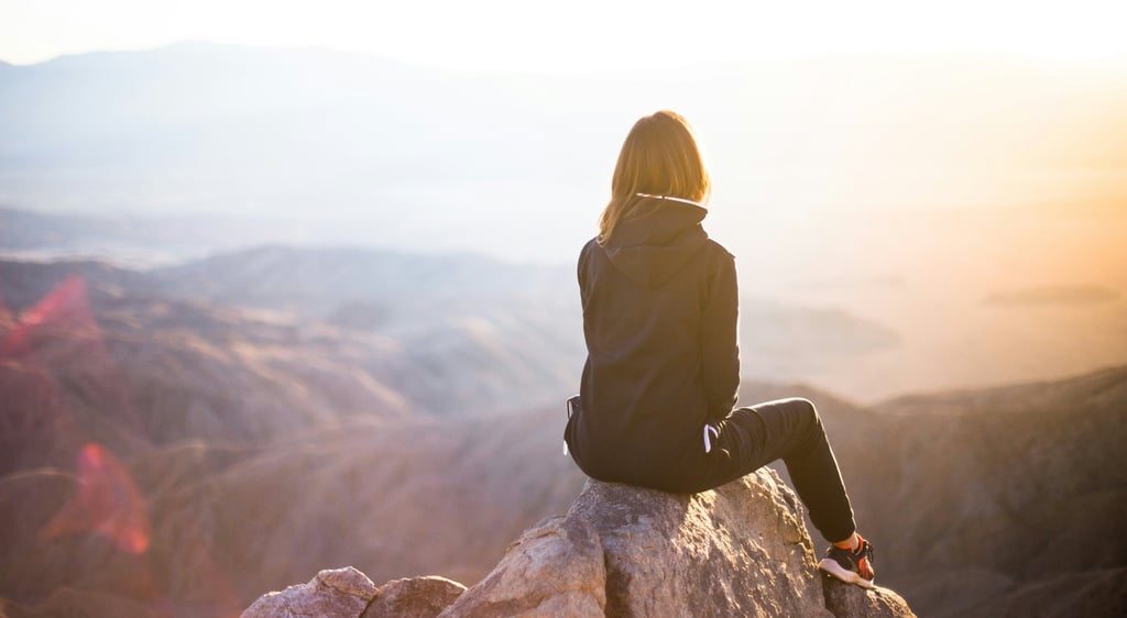 Woman sitting on mountain peak at sunrise, reflecting in solitude and enjoying peaceful nature view.