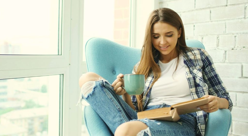 Smiling woman relaxing in a blue armchair reading a book and drinking coffee near a window.