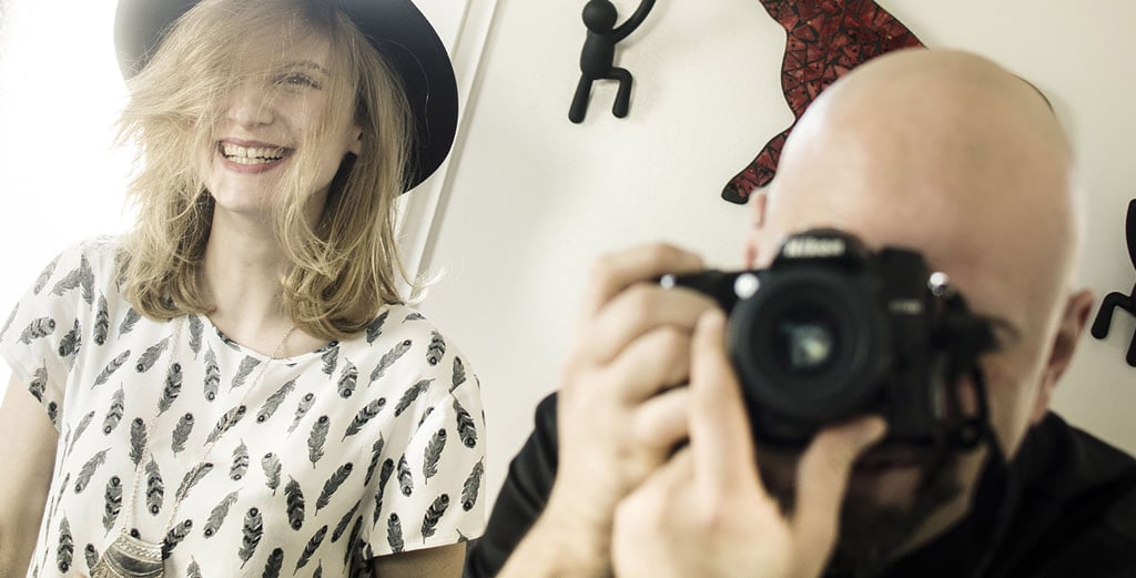A photographer, Séb taking a picture of a smiling woman wearing a black hat and feather print shirt.
