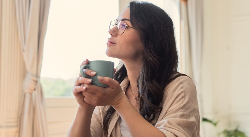 A thoughtful woman with glasses holding a coffee mug while looking out a window.