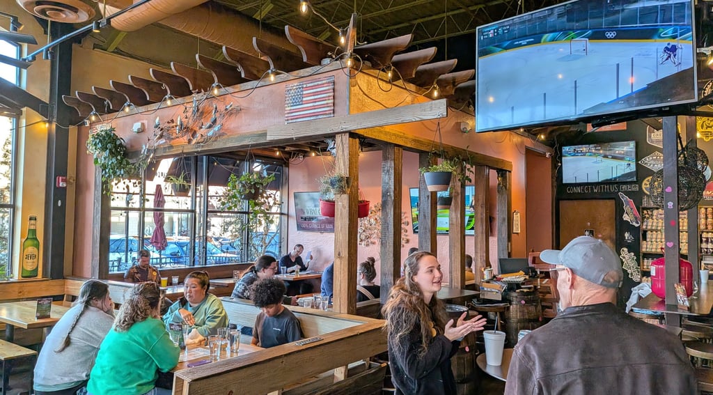 A cozy restaurant interior with customers dining at wood tables near sports TVs and hanging plants.