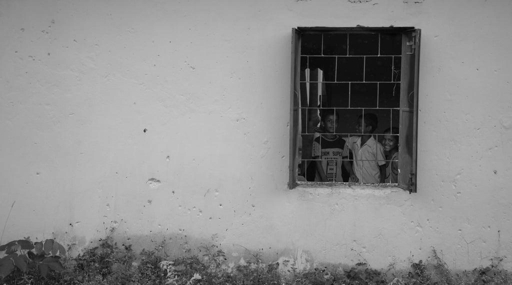 Black and white photo of three smiling children looking out of a metal barred window in a white wall.