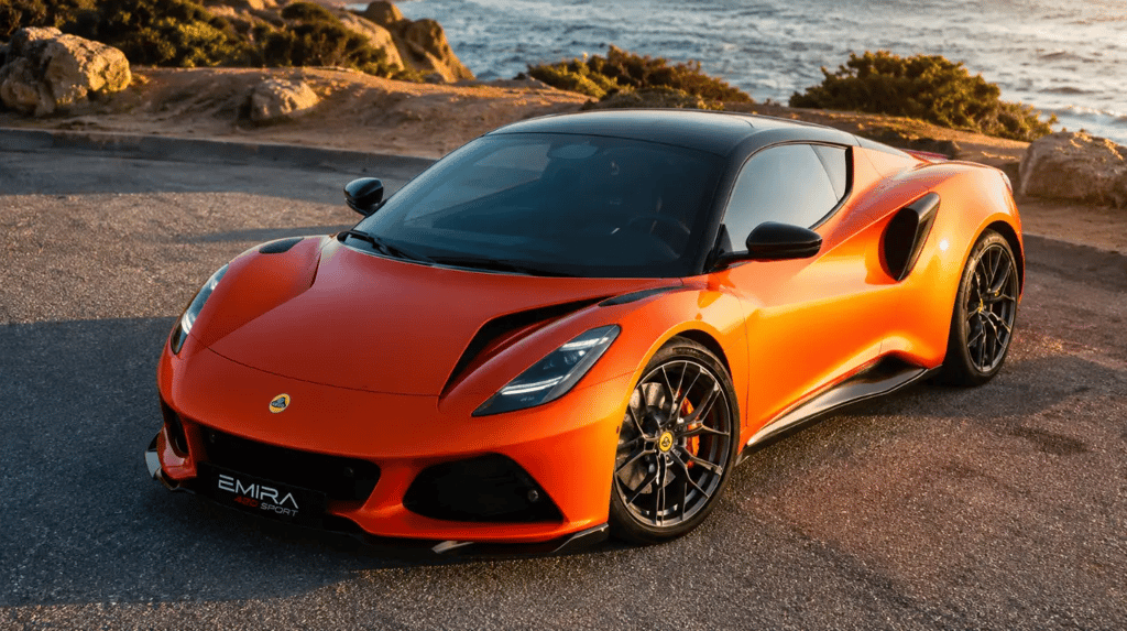 Lotus Emira 420 Sport laranja parado em estrada costeira ao pôr do sol.