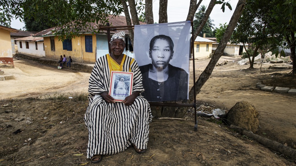 Idosa sentado ao ar livre segurando fotografia antiga, ao lado de retrato ampliado em preto