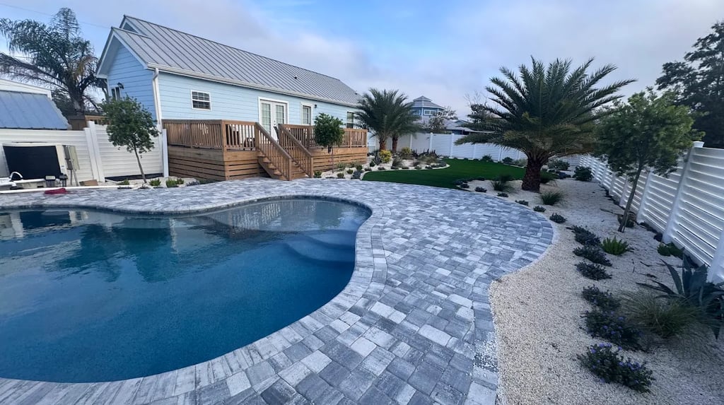 Luxury backyard with a custom swimming pool, grey paver patio, wooden deck, and tropical landscaping.