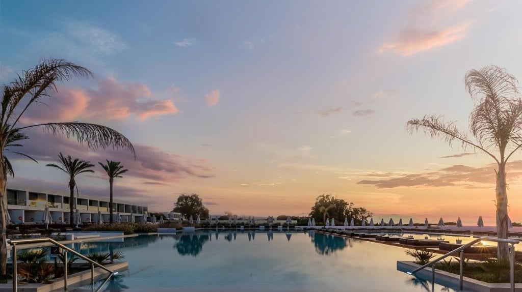 Luxury resort swimming pool reflecting a vibrant sunset sky with palm trees and lounge chairs.