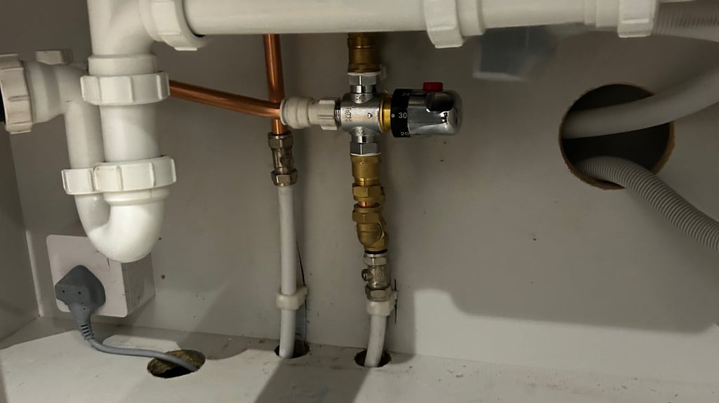 Under-sink plumbing with white PVC drain pipes, copper lines, and a thermostatic mixing valve.