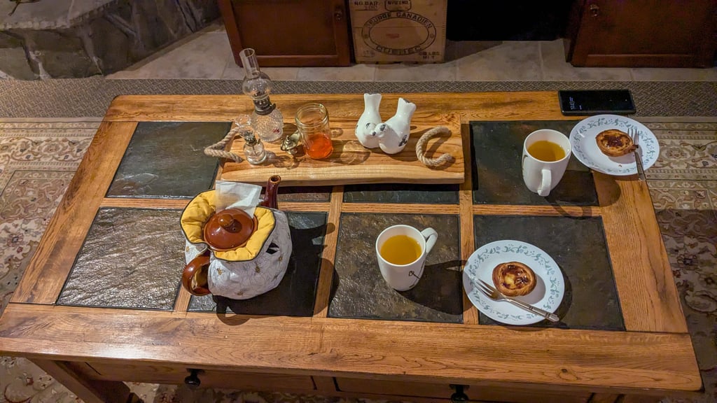 Teapot in a fabric warmer with two cups of tea and pastel de nata on a wooden table.