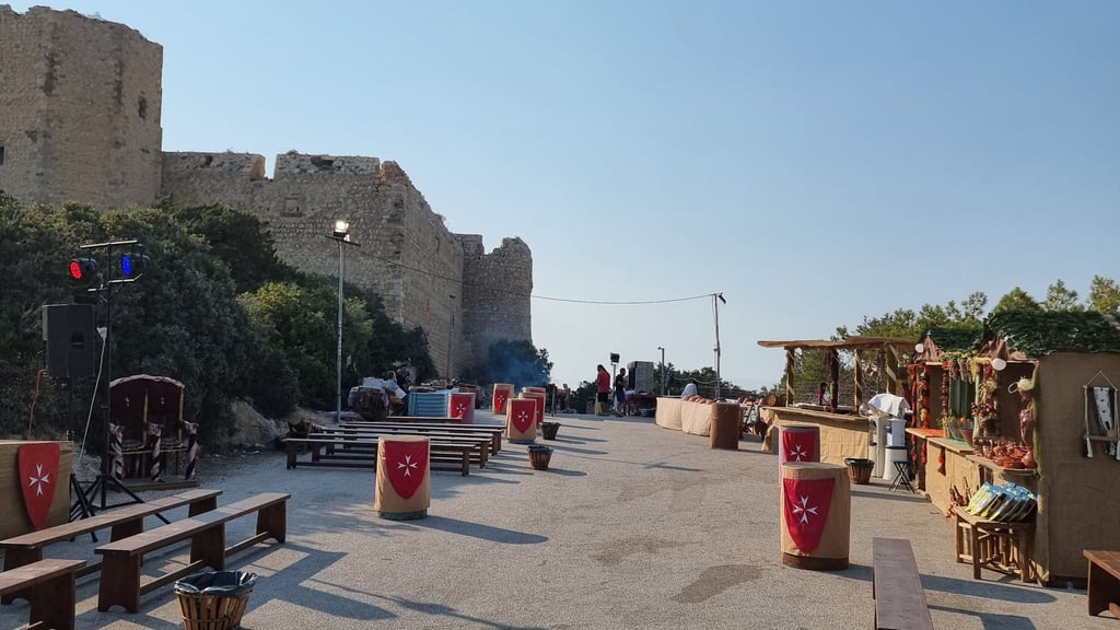 Medieval festival marketplace with wooden benches and shields near an ancient stone castle wall.