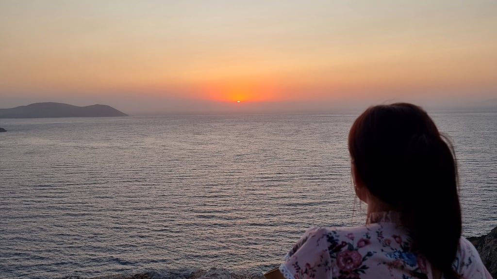 A woman in a floral shirt watches a peaceful ocean sunset from a rocky cliffside view.