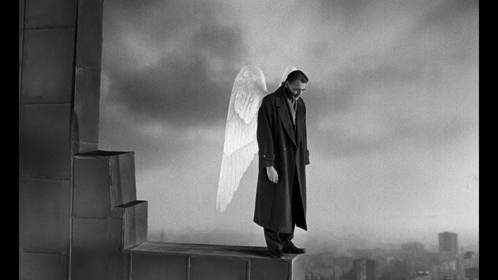 A melancholic angel with white wings stands on a rooftop ledge overlooking Berlin in a black and white film still.