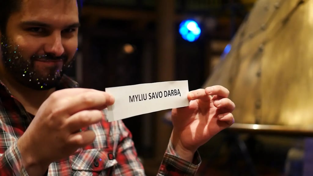 Vyras su blizgančia barzda, įmonės vakarėlyje, laiko lapą su užrašu „MYLIU SAVO DARBĄ"