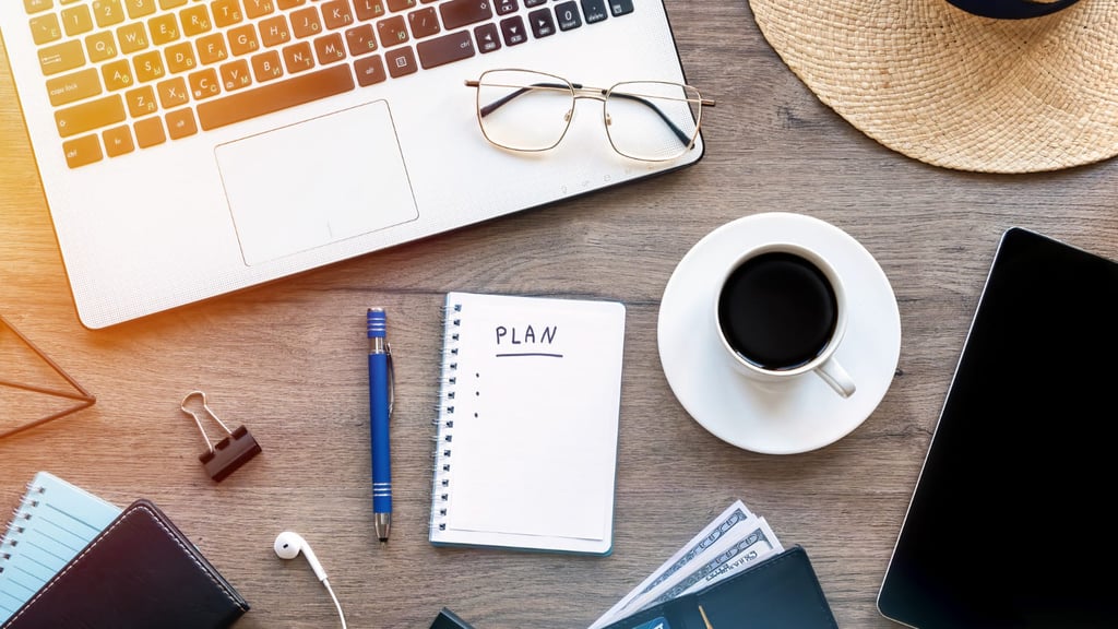 Uma mesa com notebook, xícara de café, caderneta, óculos, lapiseira, fone de ouvido e brochura.