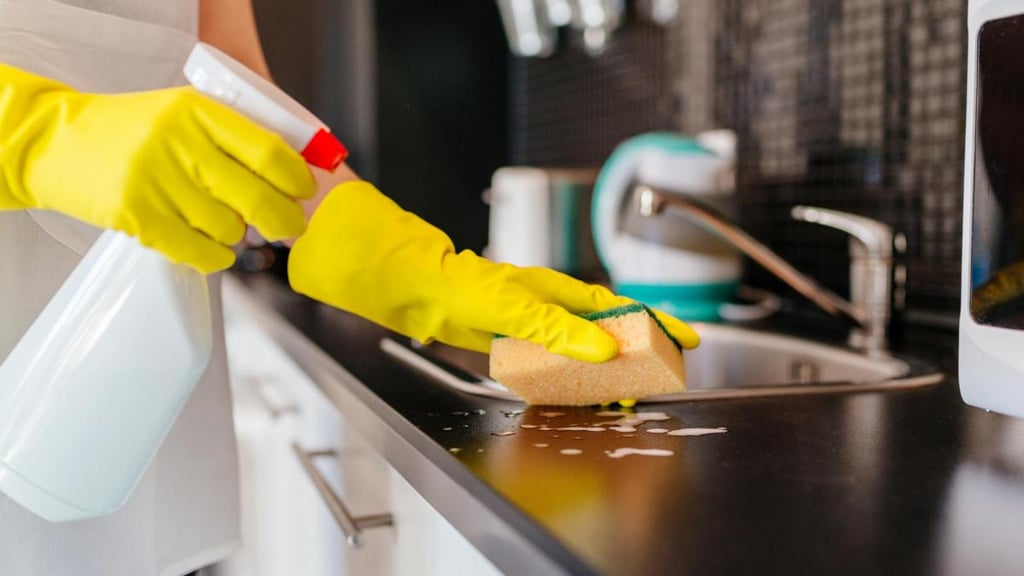 A person wearing yellow rubber gloves uses a sponge and spray cleaner to sanitize a kitchen countertop.
