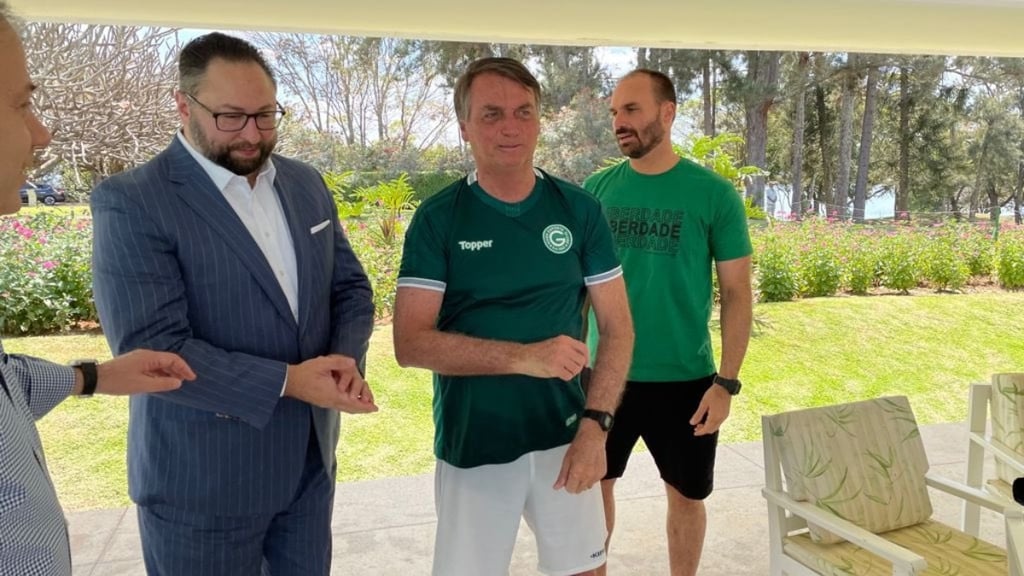 Jair Bolsonaro em pé, com camisa de futebol do Goiás, ao lado de dois homens.