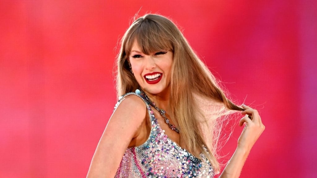 Taylor Swift sorrindo, com um vestido brilhante e puxando o cabelo, em um show.