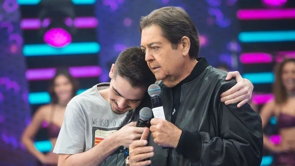 Faustão abraça e segura um microfone com seu filho com uma camisa cinza