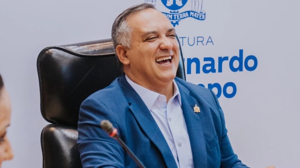 em um terno azul, sentado em uma cadeira, rindo.