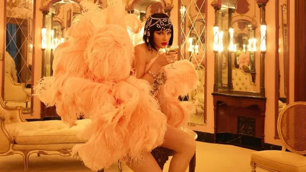 Taylor vestindo penas cor-de-rosa, com um copo de champanhe, em um quarto luxuoso.