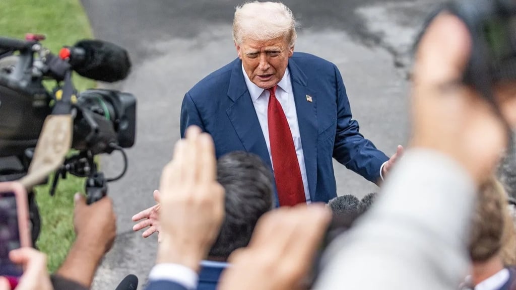 Donald Trump, com terno azul e gravata vermelha, em meio a repórteres e câmeras.