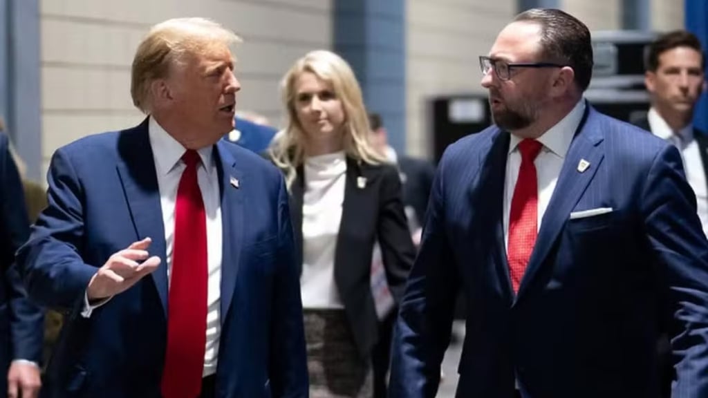Donald Trump caminha e conversa com um homem de terno, com outras pessoas ao fundo.