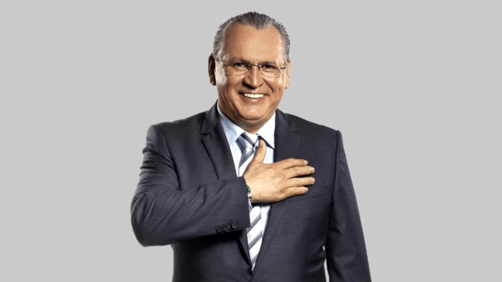 Sidney Oliveira sorrindo em um terno azul, com a mão direita sobre o peito.