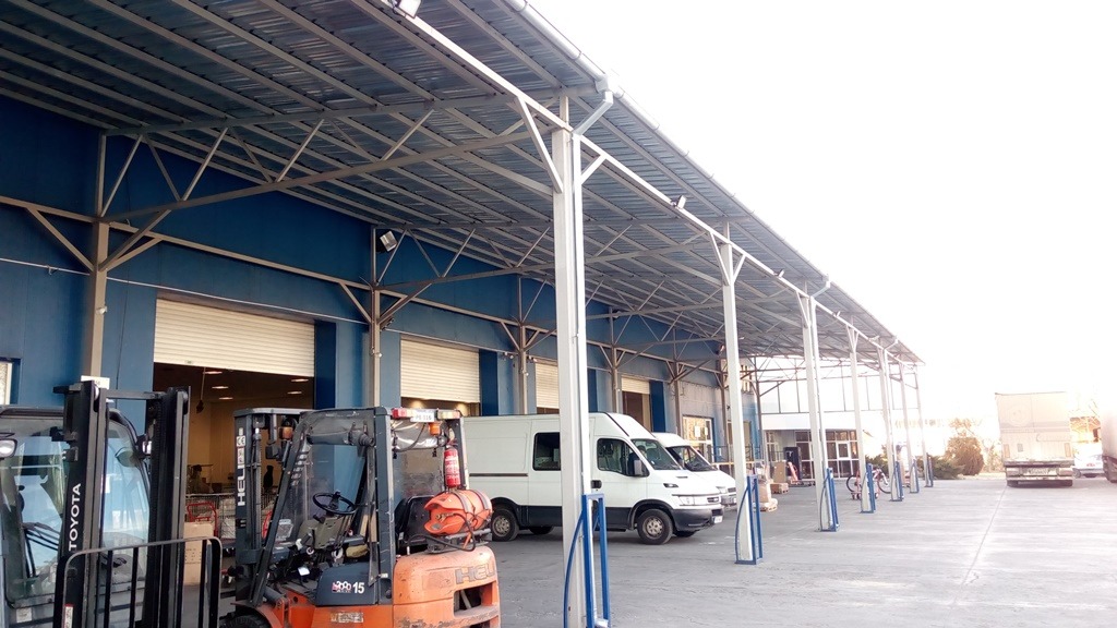 Logistics warehouse loading dock with parked forklifts, delivery vans, and a metal canopy.