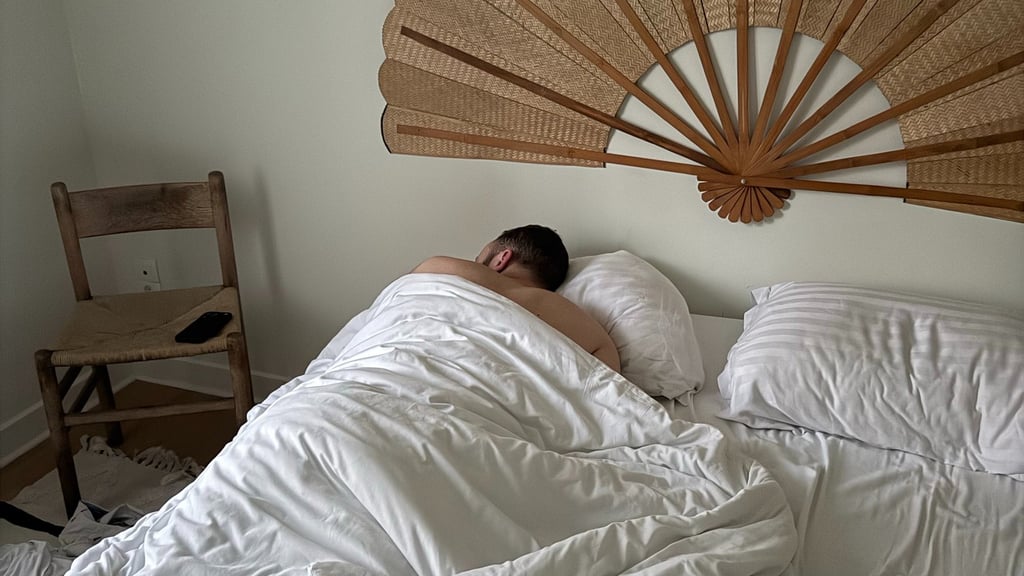 A man sleeping in a bed with white linens below a large decorative rattan fan headboard.