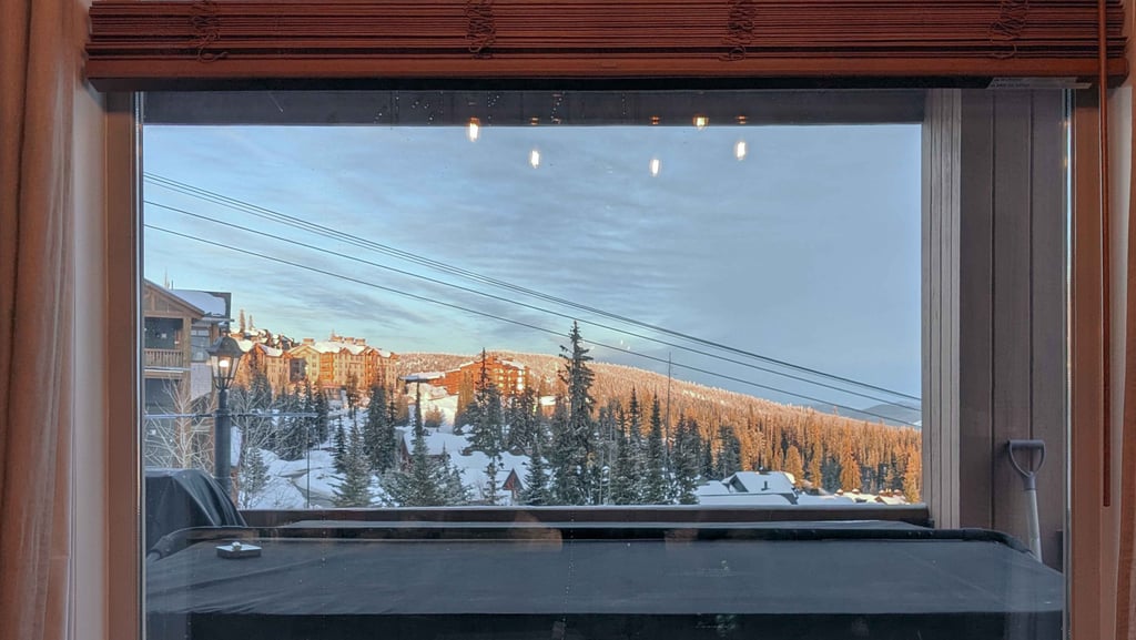 Snow-covered trees and mountain buildings seen through a window at sunset.