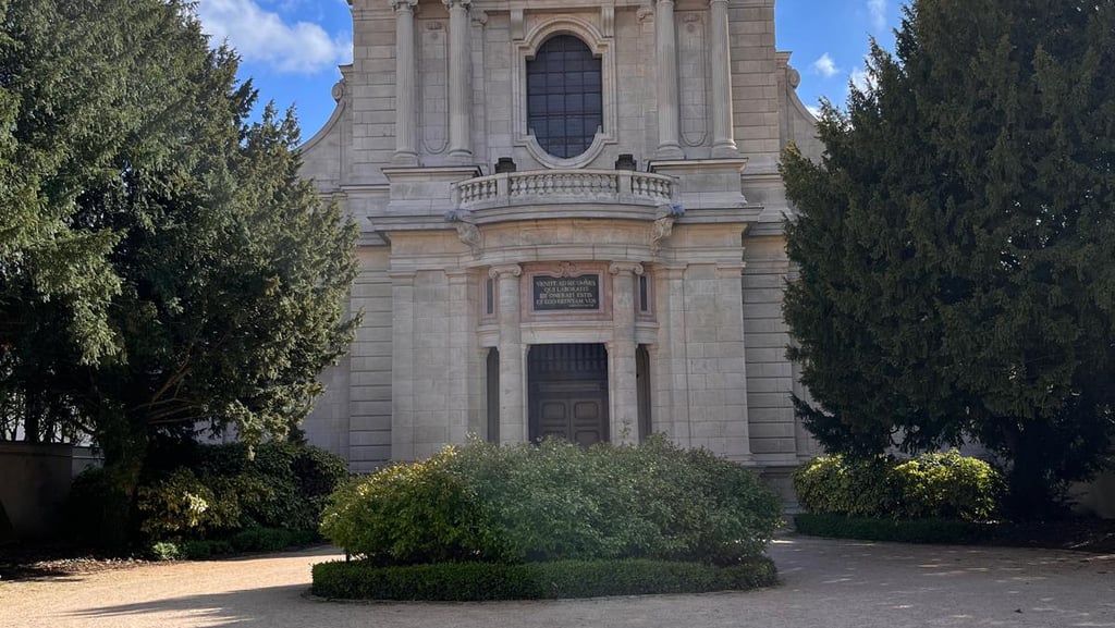 Historic stone chapel with classical architecture, green trees, and a gravel courtyard under a blue sky.