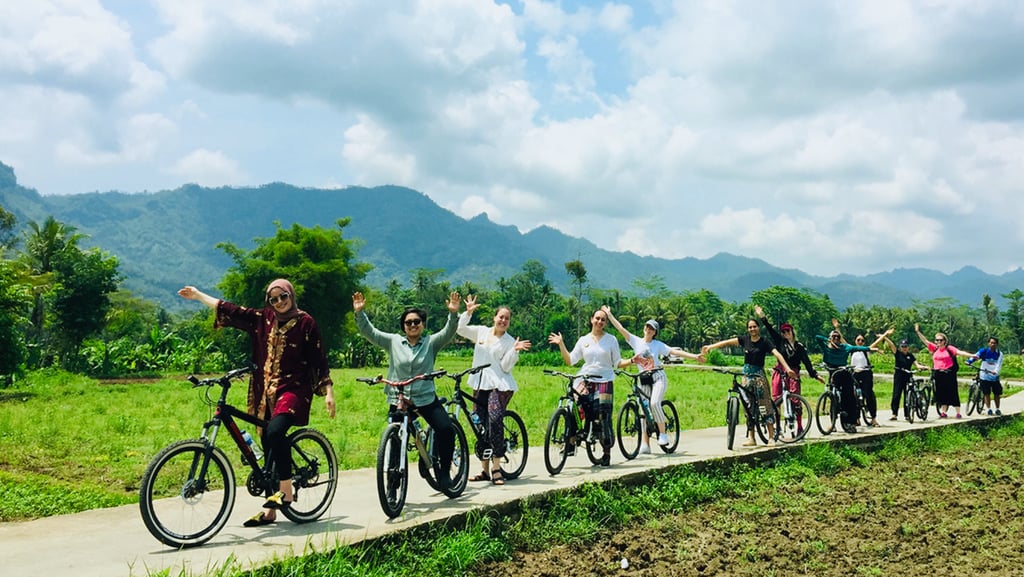 Wisatawan bersepeda santai menyusuri jalan pedesaan dengan pemandangan sawah dan Candi Borobudur di kejauhan