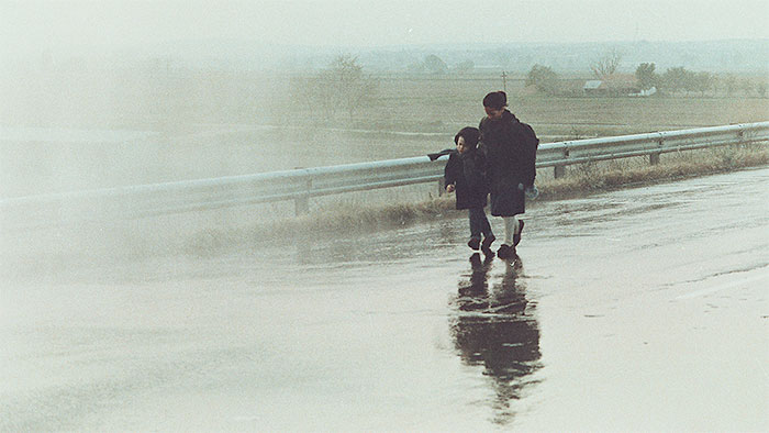 A wide, misty shot of two people walking along a wet, reflective roadside under a hazy grey sky.