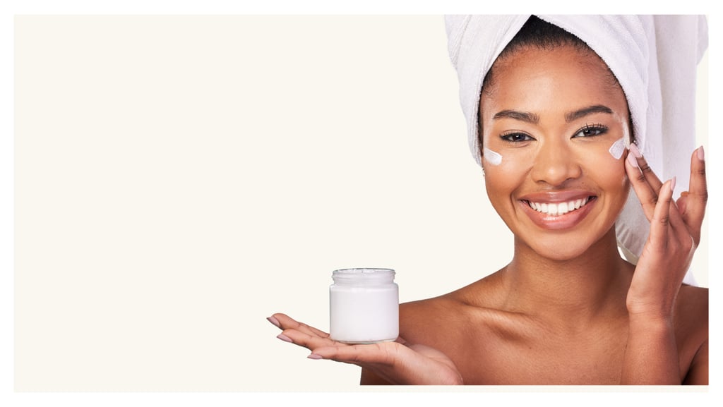 Smiling woman with a towel head wrap applying moisturizing face cream from a white skincare jar.