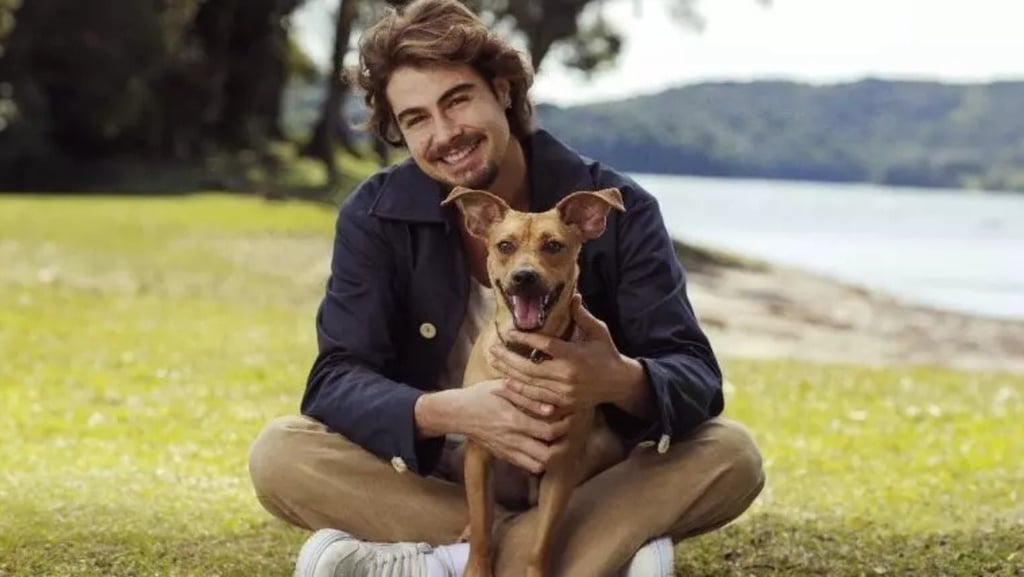 Rafael Vitti sorri, sentado na grama, abraçando um cachorro viralata caramelo.