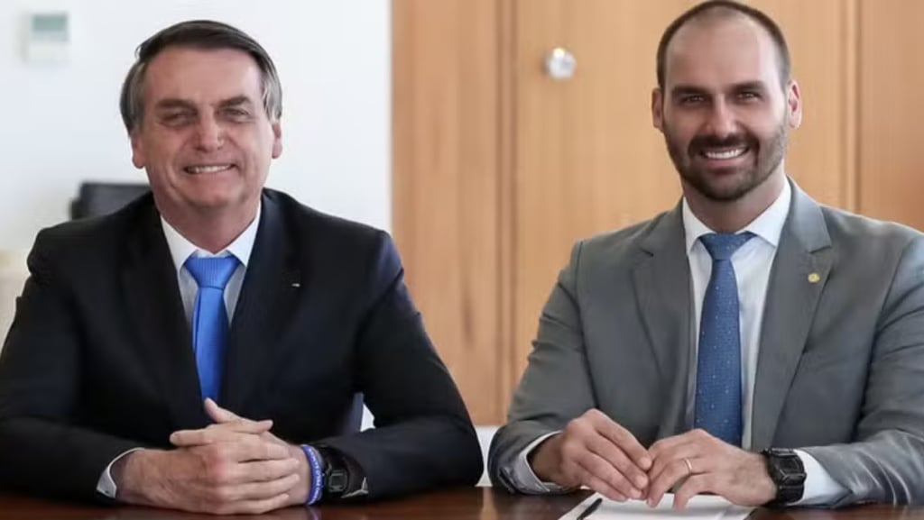 Jair Bolsonaro e Eduardo Bolsonaro sentados em uma mesa, ambos sorrindo.