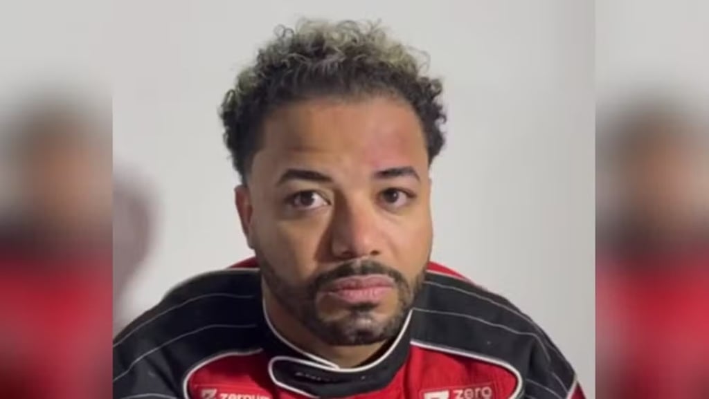 Hytalo Santos com cabelo loiro, usando um macacão de corrida vermelho.