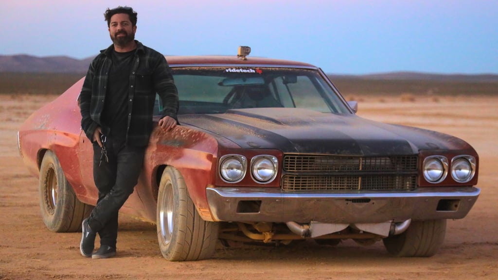 A man leans against a classic red and black muscle car in the desert at sunset.