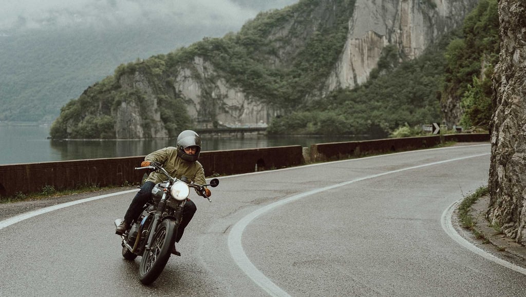 A person riding a vintage motorcycle on a winding coastal road with scenic mountain views.