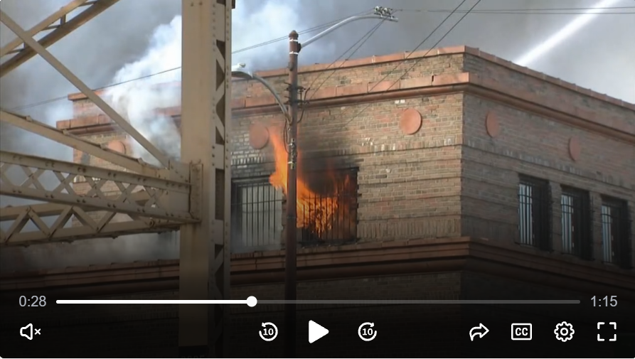 Flames and smoke pour out of a brick building window during a structure fire with active firefighting efforts.