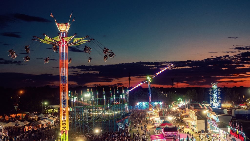 Aerial view of a vibrant night carnival with illuminated amusement park rides, games, and crowds at sunset.