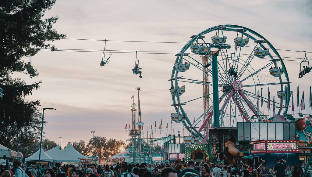 Crowds at a summer carnival with a Ferris wheel, chairlift ride, and ticket booths at sunset.
