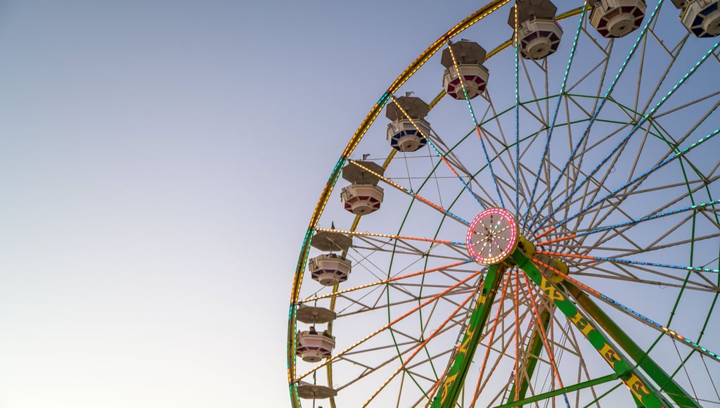 A large Ferris wheel with colorful LED lights spinning against a clear twilight sky at a local carnival.