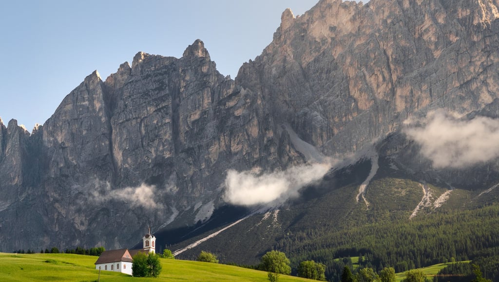 A small white chapel sits on a lush green meadow beneath the jagged peaks of the Italian Dolomites.