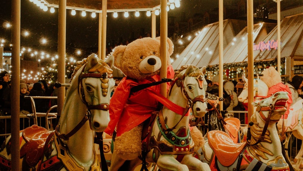 A large teddy bear in a red coat riding a vintage carousel horse at a festive outdoor carnival.