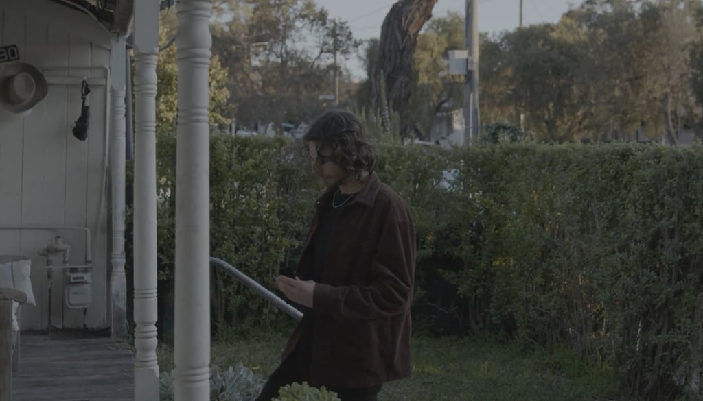 A man with long hair wearing a brown jacket stands on a porch next to a white pillar and green hedge