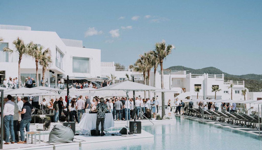 Guests enjoying a luxury beach club pool party with a DJ and white Mediterranean architecture.