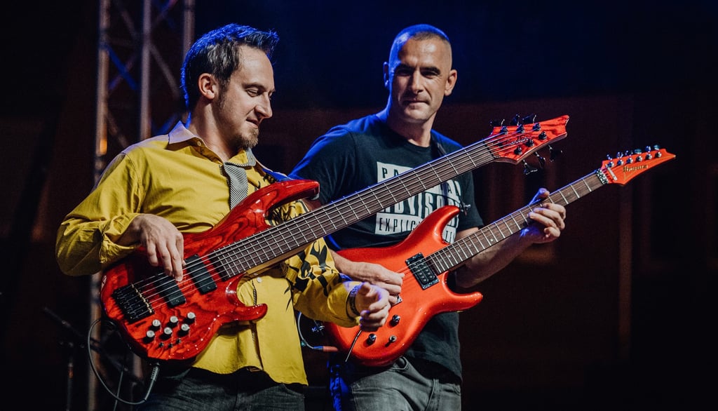 Two guitarists performing on stage with matching red guitars under blue stage lights.