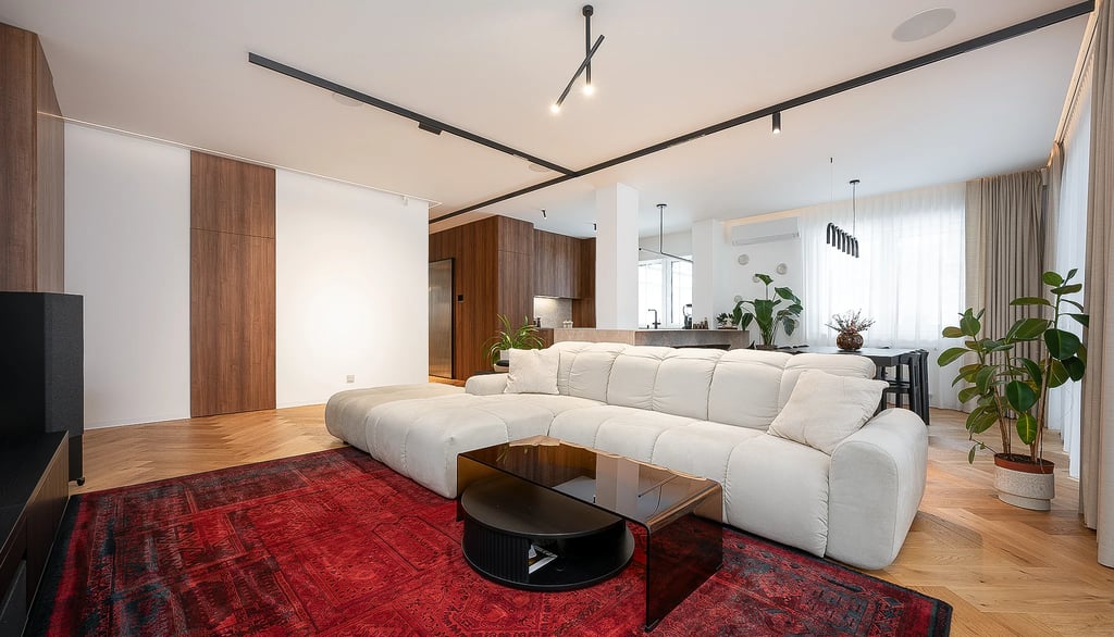 Nowoczesny salon z jasną sofą narożną, czerwonym dywanem, stolikiem i aneksem kuchennym.