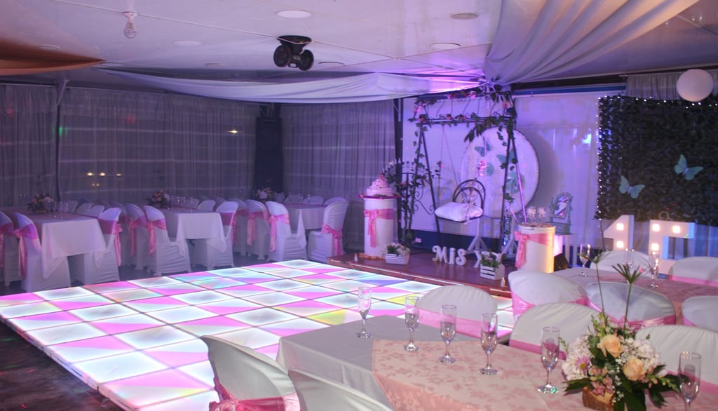 Salón elegante con pista de baile iluminada, mesas decoradas y detalles florales.